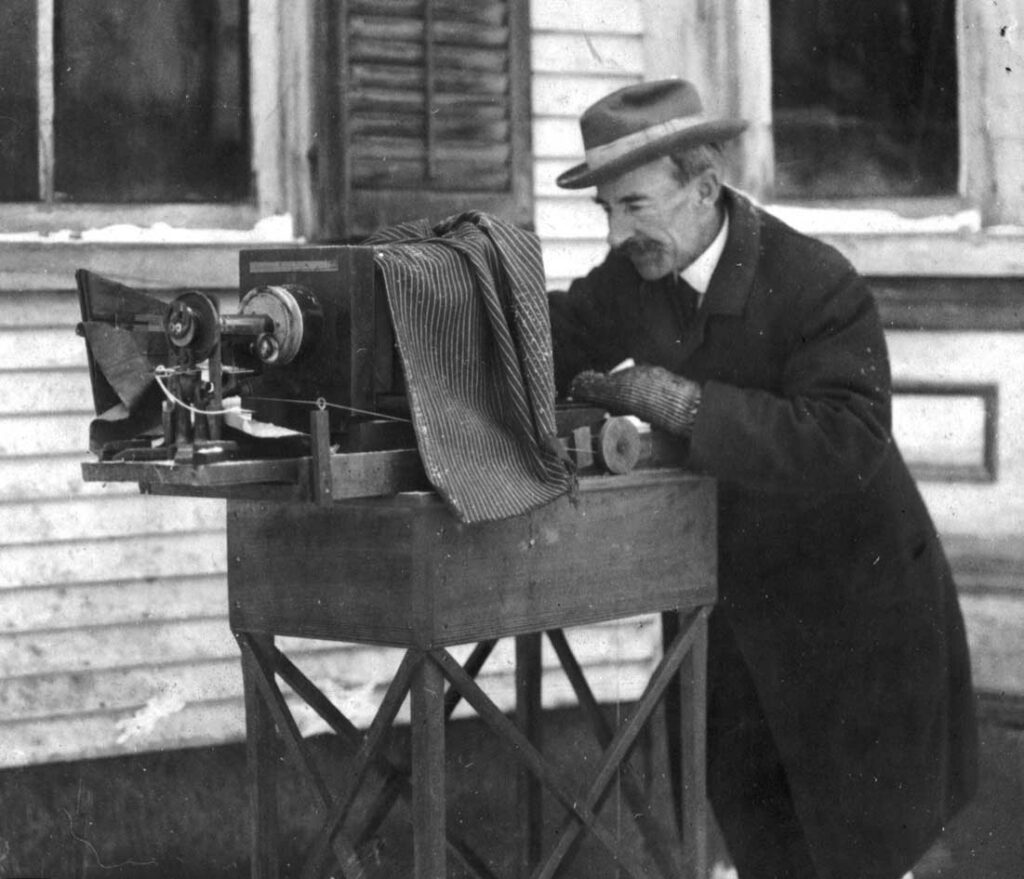 Black and white photo of an older man outdoors in a coat and hat standing behind a camera
