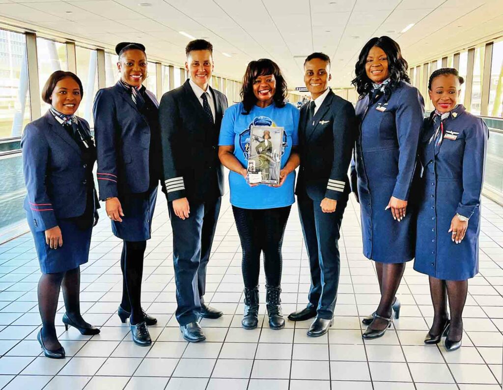 A woman holds a Bessie Coleman doll and poses with six women in airline uniforms.
