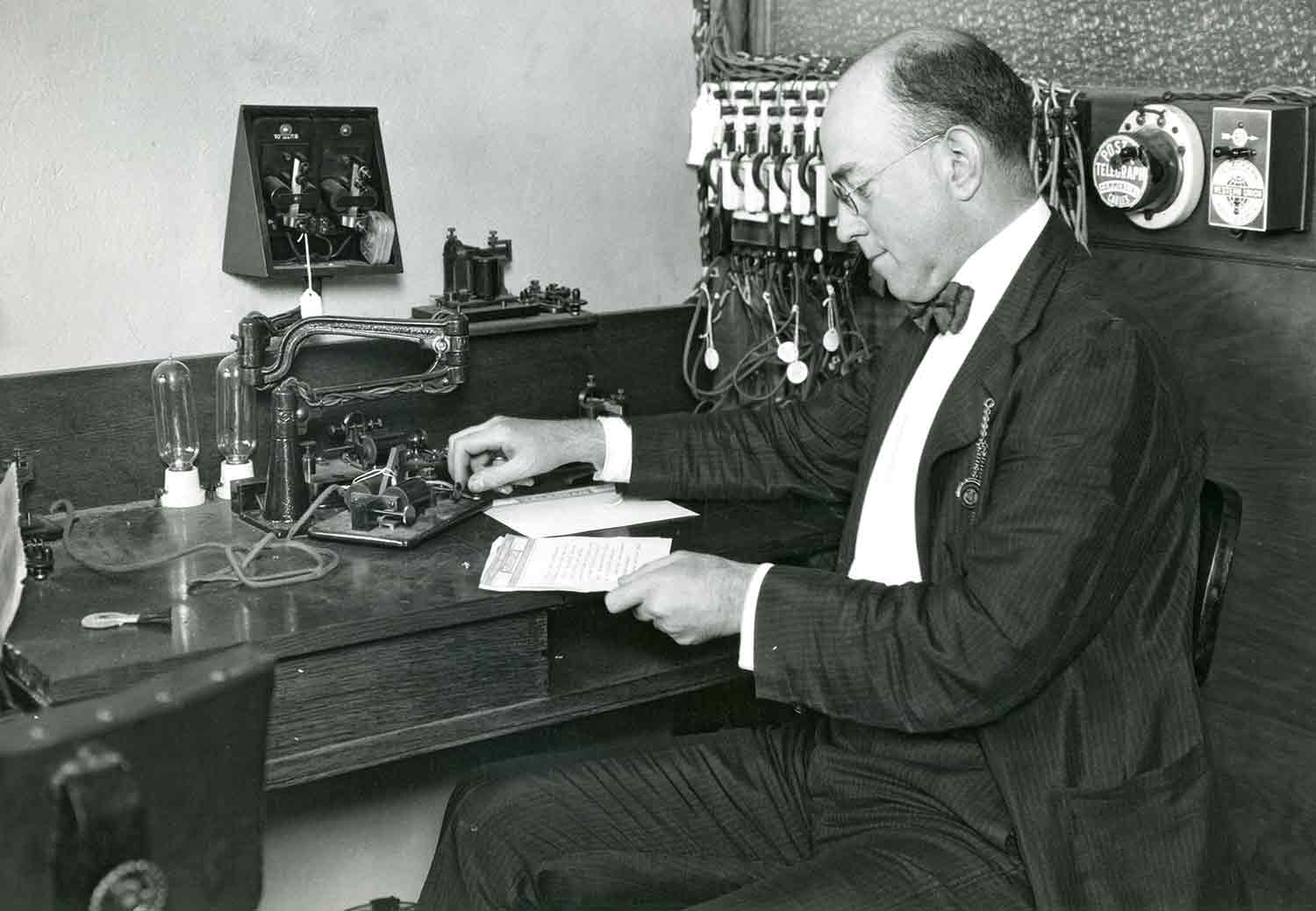 A man in a suit and bowtie looks at a message on a piece of paper while using a telegraph machine.