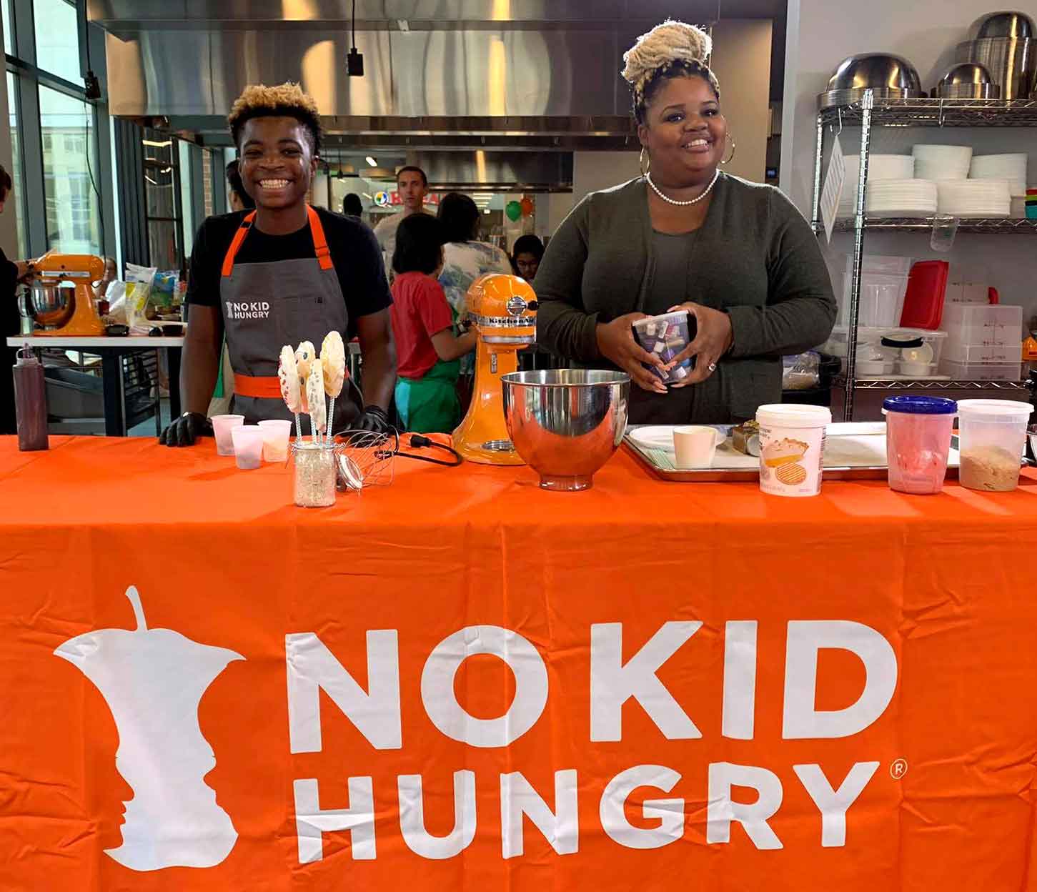 A teen and a woman stand at a table with a No Kid Hungry tablecloth and baking equipment.