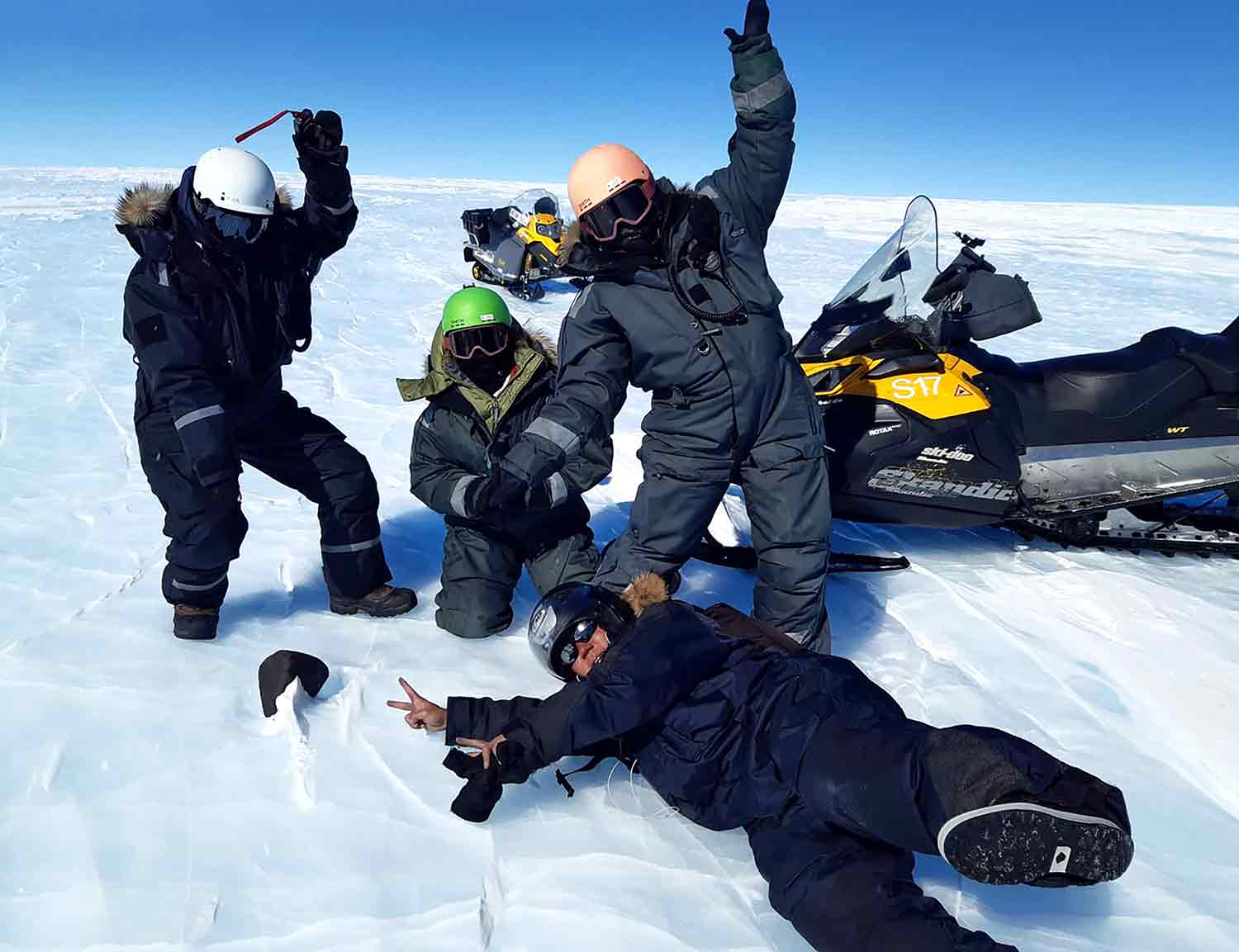 Four people in snowsuits, helmets, and sunglasses on an icy landscape gather around a meteorite.