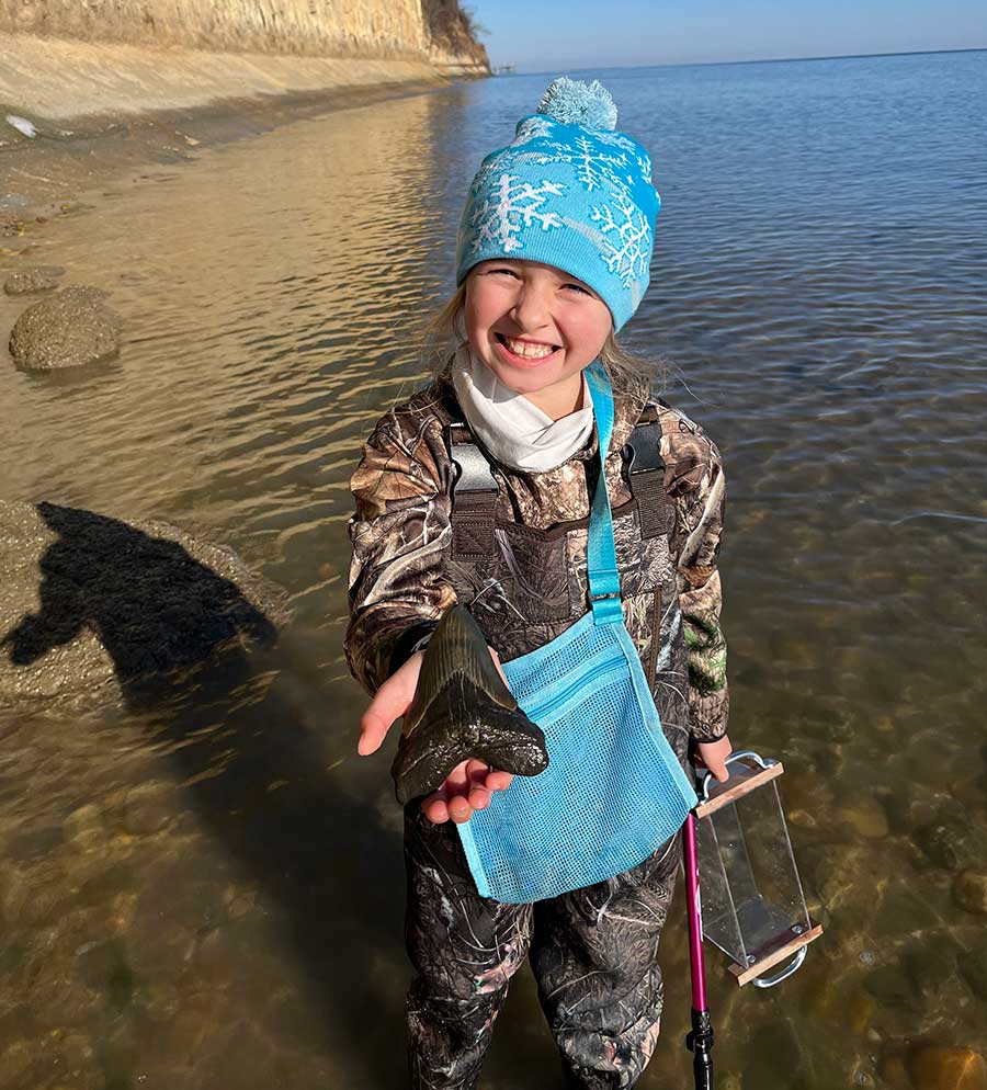 A young child stands in shallow water and holds up a large fossil of a shark tooth.