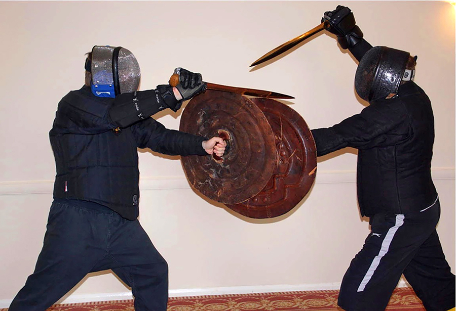 Two people wearing face shields and holding metal shields fight with bronze swords.