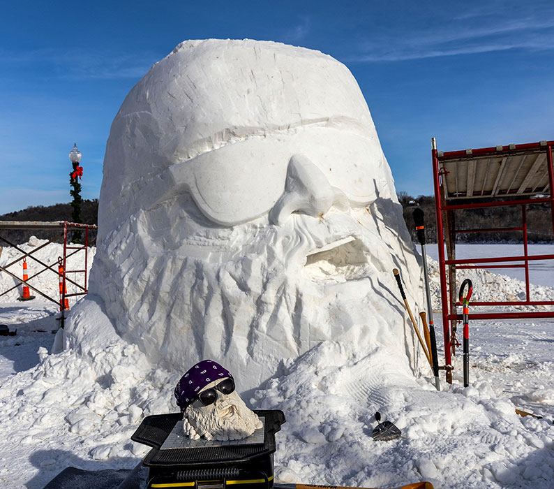 A large snow sculpture of a bearded biker in sunglasses with smaller clay model sitting in front.