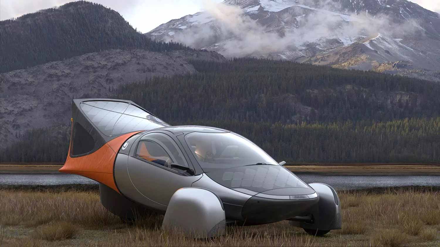A three-wheeled black and silver car with a tent mounted in the rear sits in a field with a stream and mountains in the background.