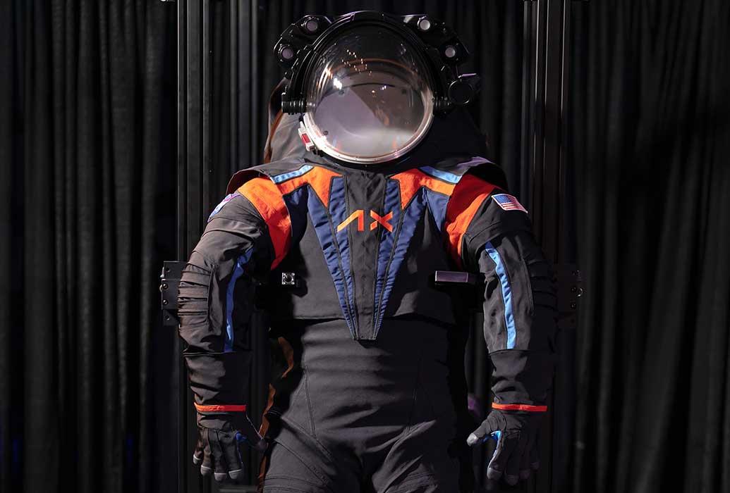 A black helmet and space suit with orange and blue trim on display in front of a black curtain