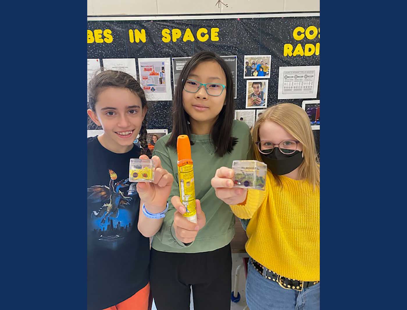 Three young people pose together, with two holding clear plastic cubes and the other holding an EpiPen.