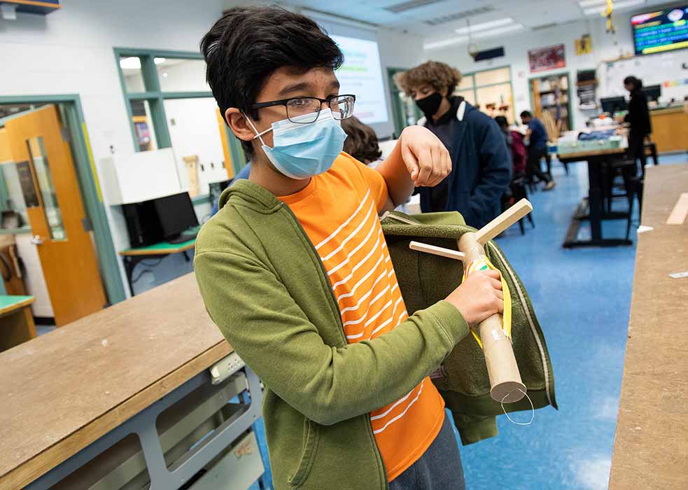 A teen uses a cardboard and wood stick to pull a zip up sweatshirt over his arm and shoulder.