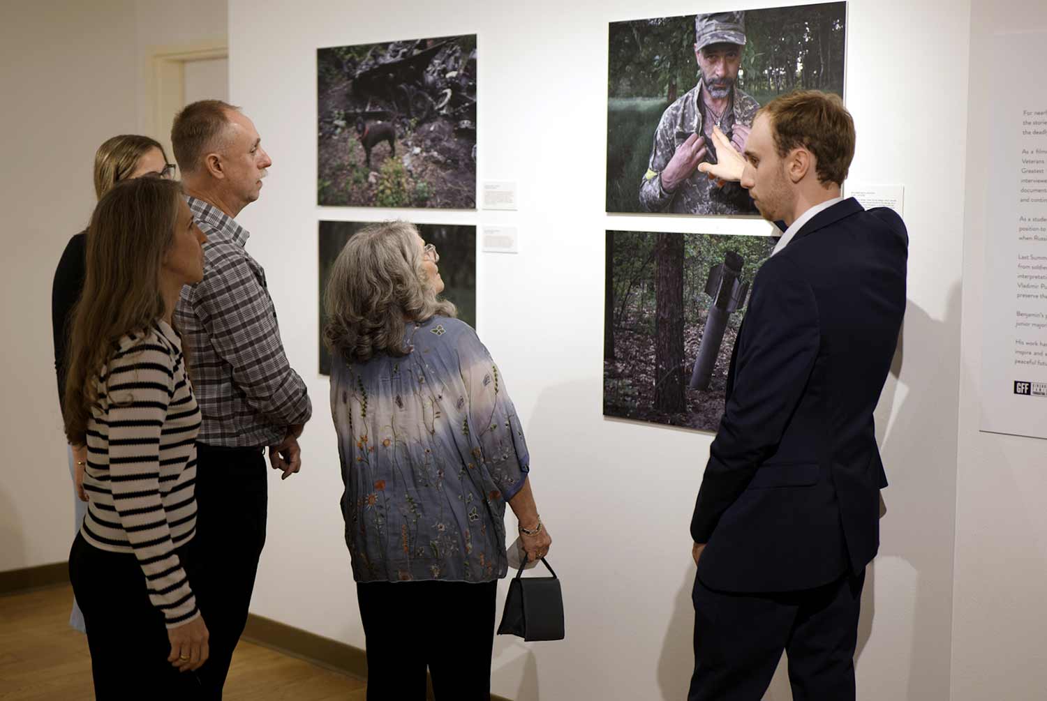 A young man points gestures to a wall of photos showing scenes from Ukraine as several people study the images.