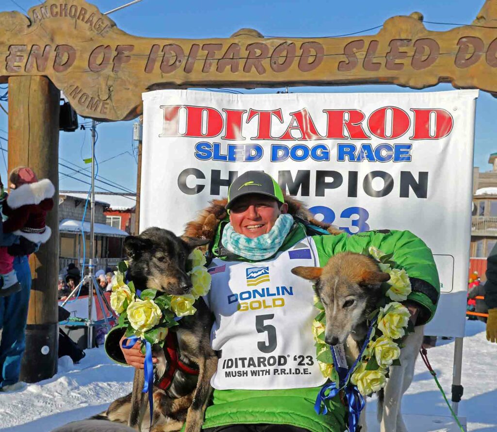 A man with each arm around a dog poses in front of a banner saying Iditarod Sled Dog Champion.