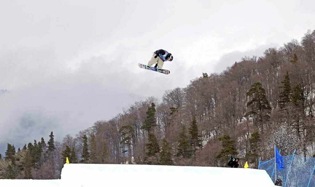 A snowboarder on a board flies high over a slope as a man takes her photo.