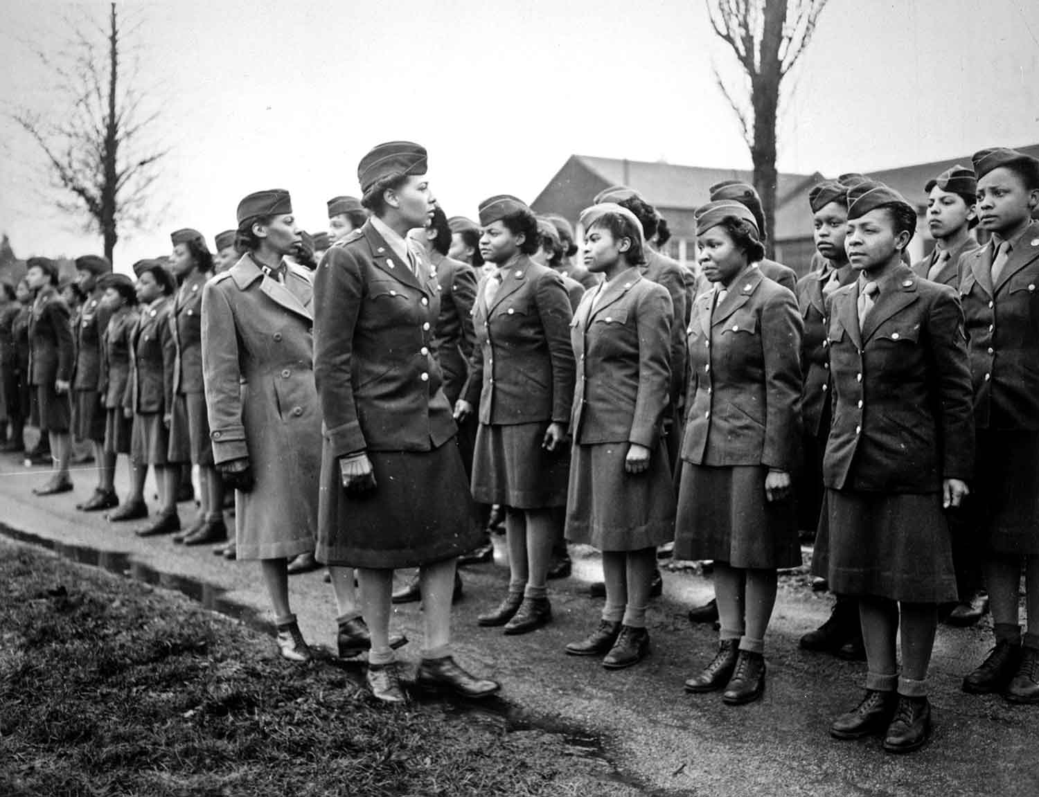 Two women in uniforms stand before a group of women lined up in rows.