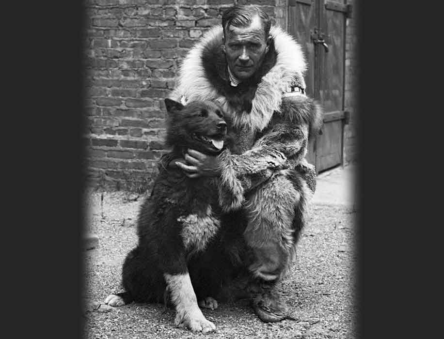 A man in a fur coat crouches on the ground with his arms around a husky-type dog.