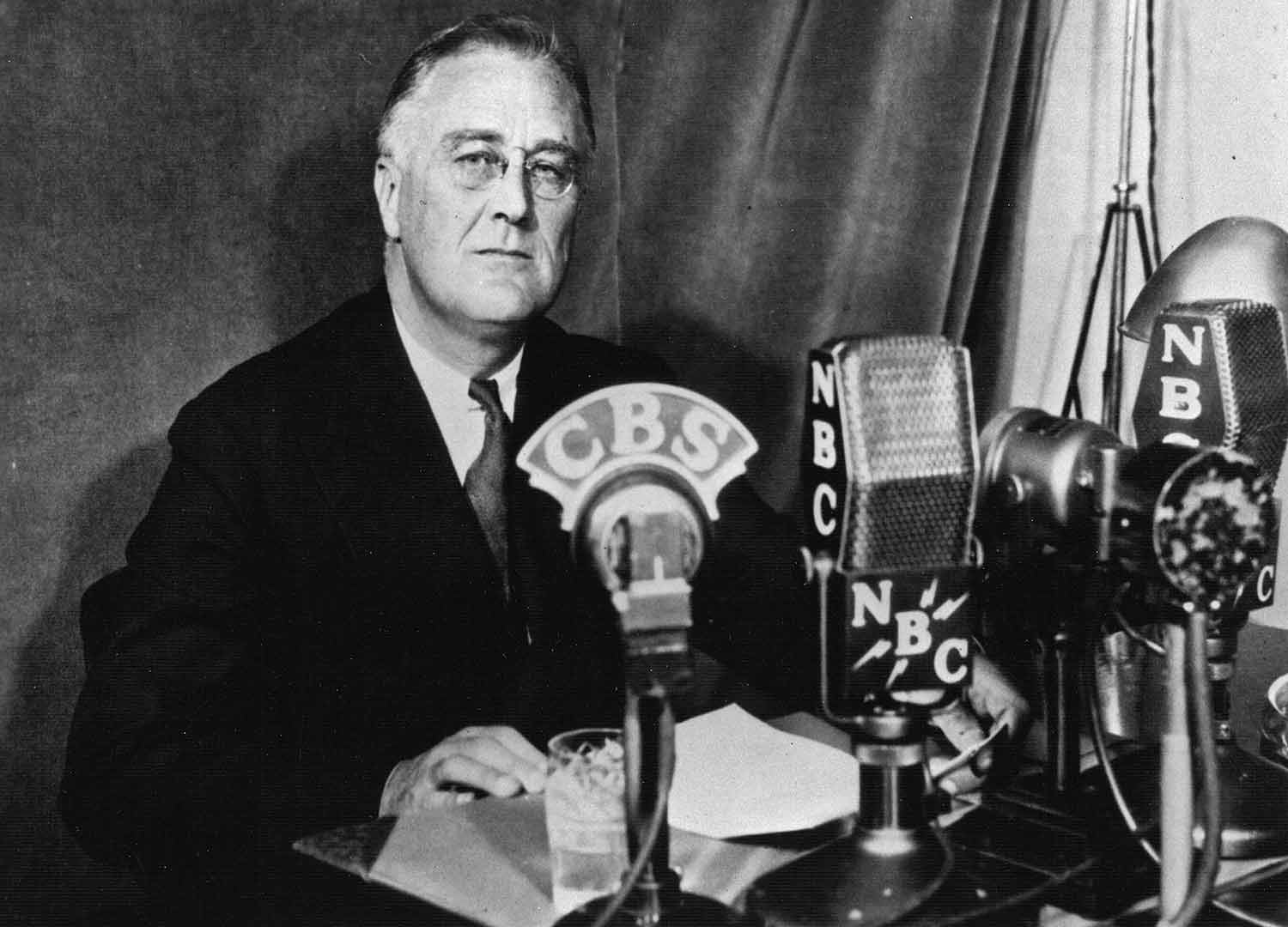 Franklin D. Roosevelt sits in front of radio microphones and looks at the camera.
