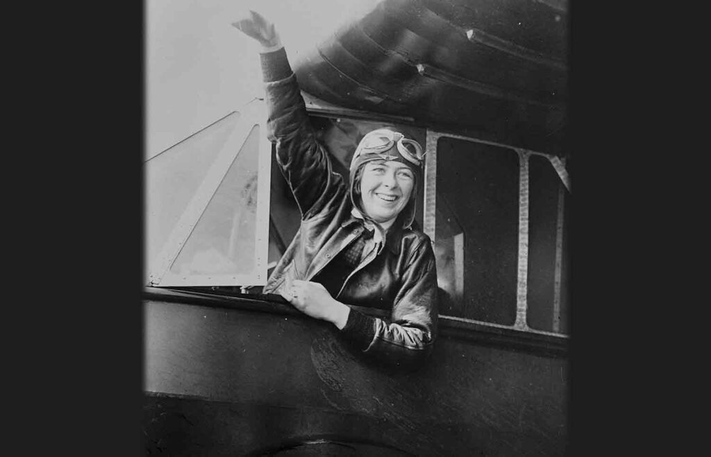 A young woman in early aviation clothes waves from the cockpit of a plane.