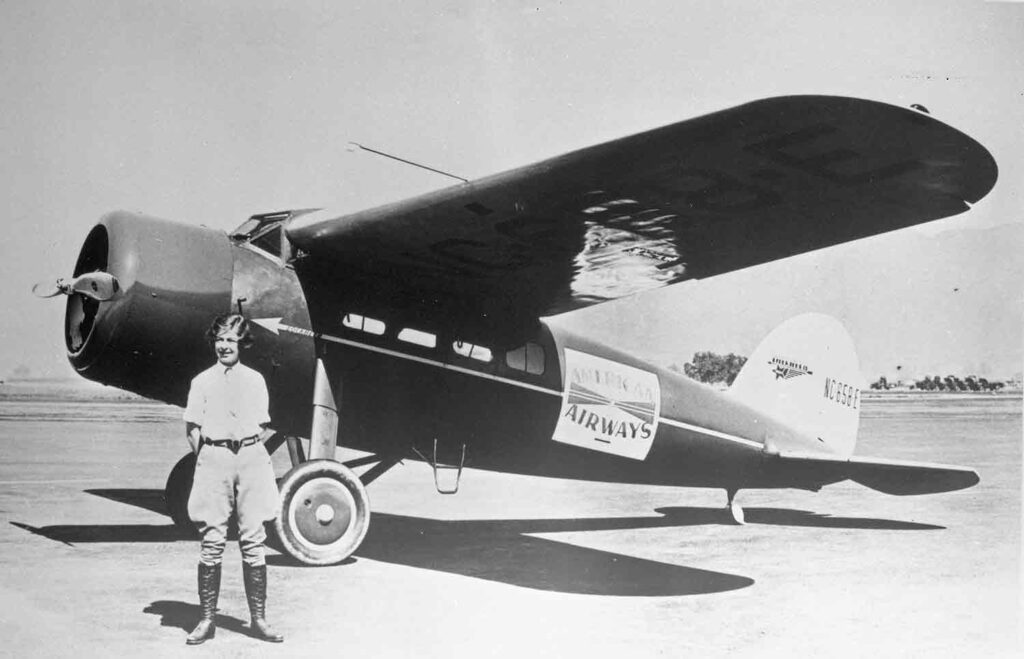 A young woman wearing breeches and boots poses in front of a small plane.