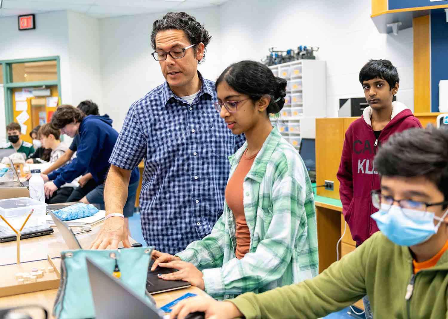 In a classroom, a teacher and student look at a computer as other students work at tables.