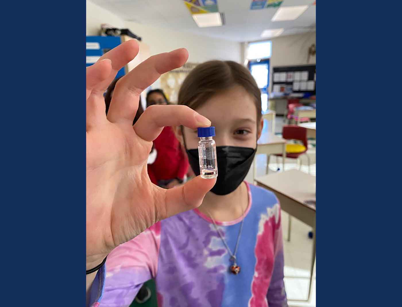 A girl in a classroom holds a vial with clear liquid inside.