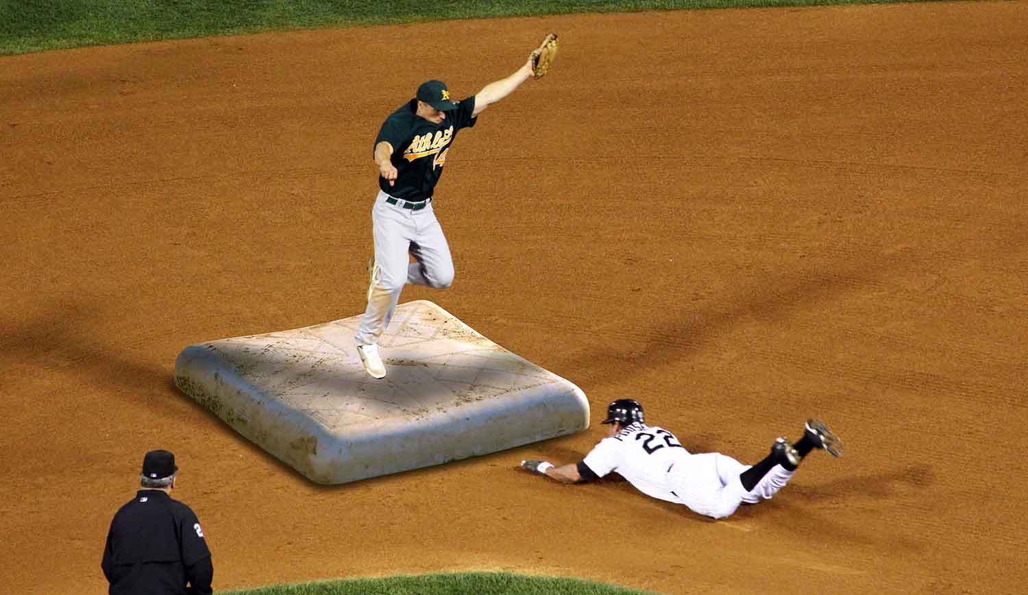 A baseman stands on a very large base catching a ball as a hitter slides toward the base.