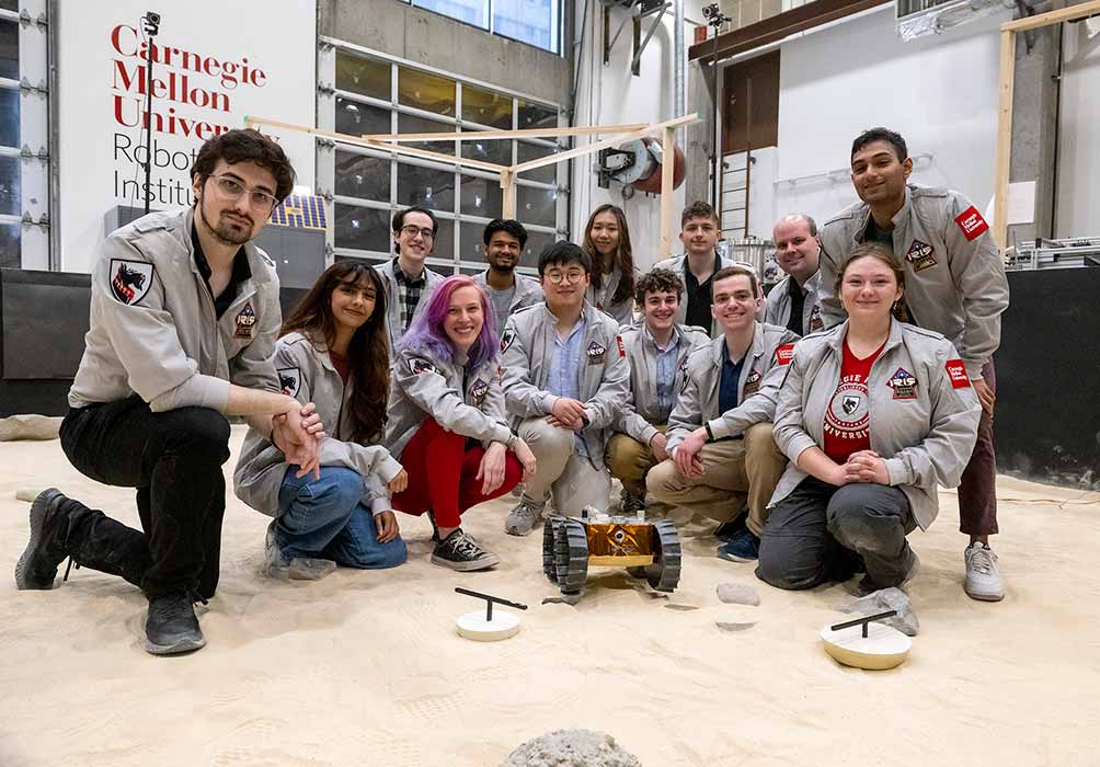 A group of young adults kneel behind a small rover on a sandy surface inside a building.