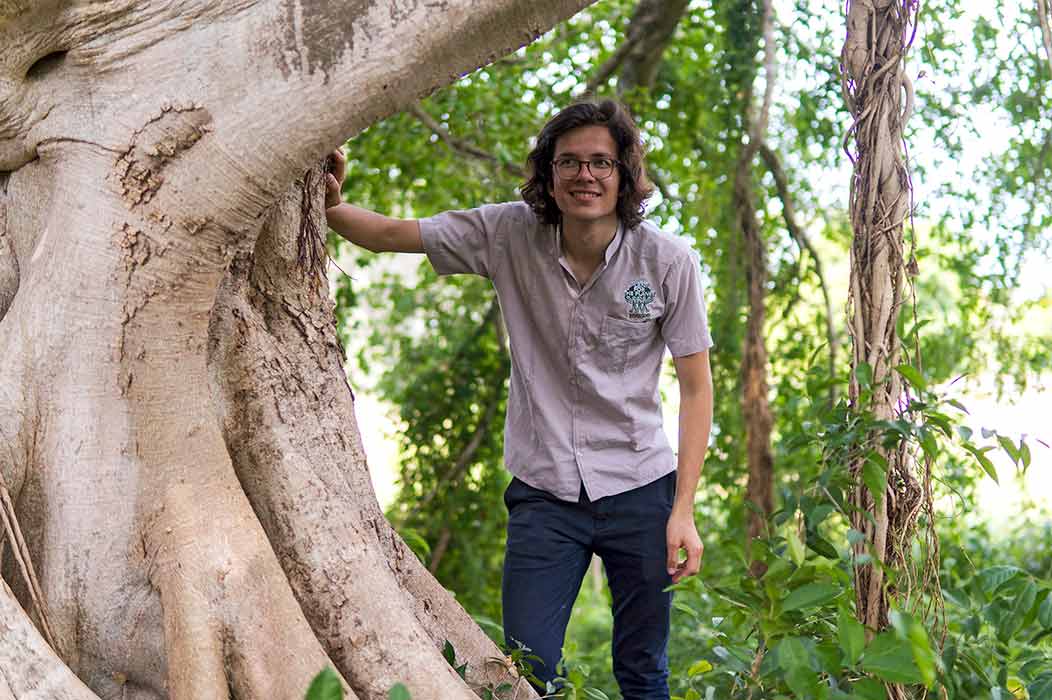 A young man leans with his hand on a large tree trunk.