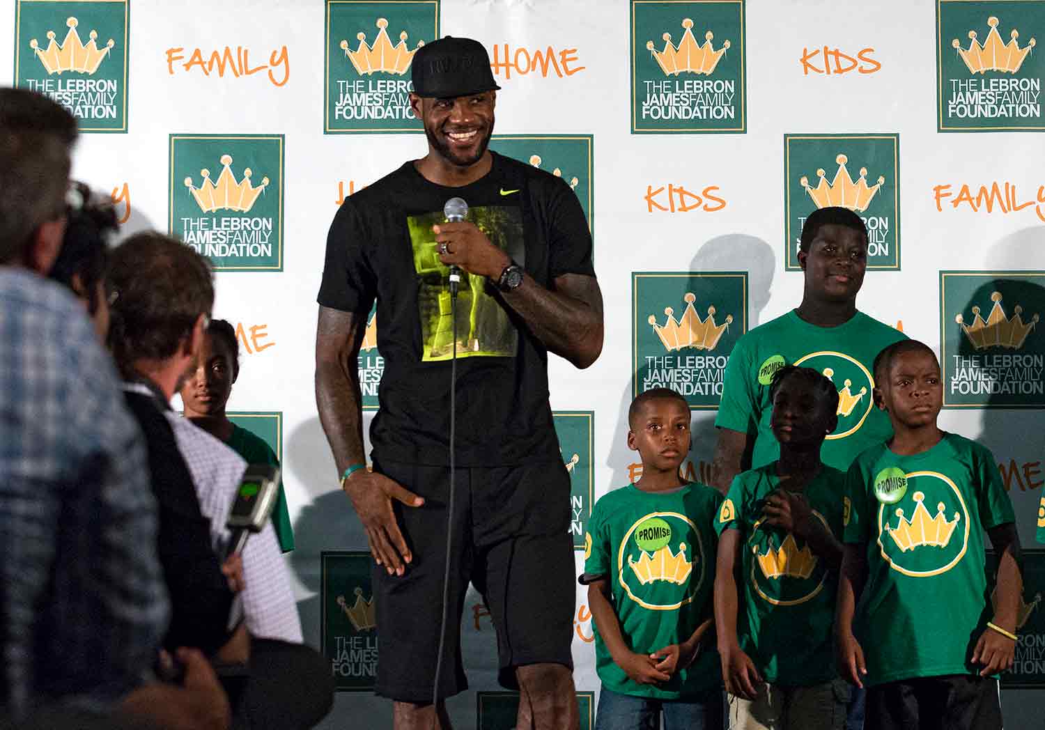 LeBron James smiles while holding a microphone and standing next to children in front of a small audience.