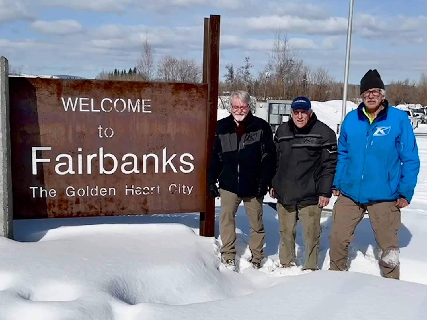 Three men pose next to a sign that says Welcome to Fairbanks, the Golden Heart City.