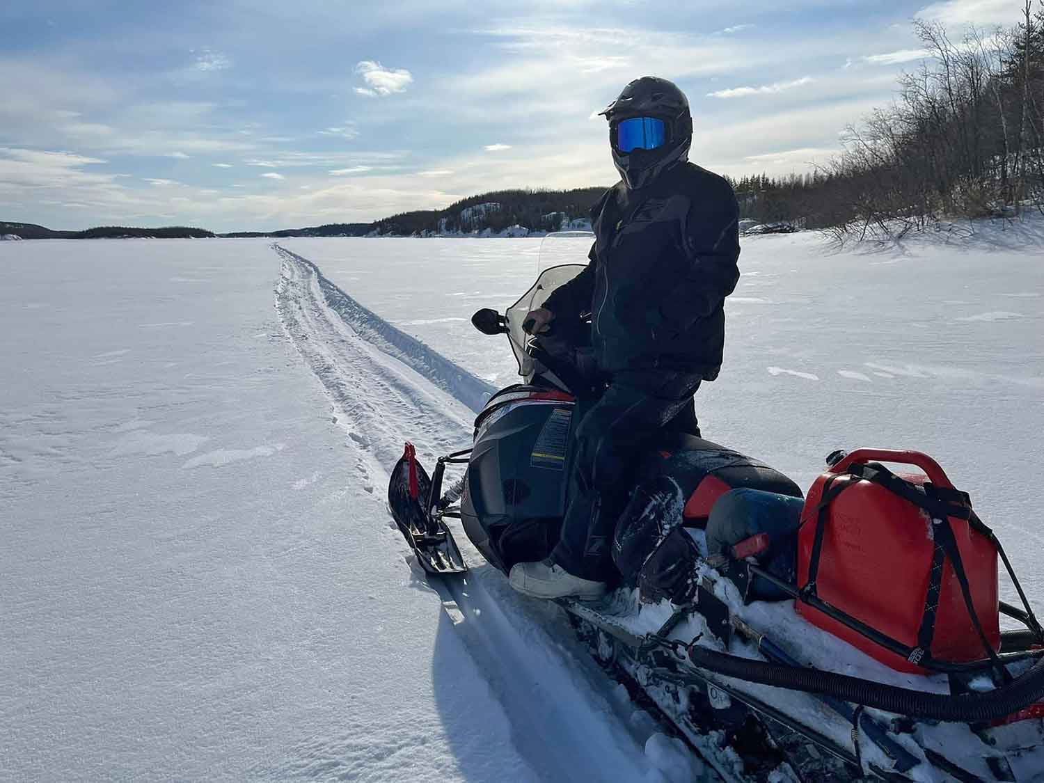 A man on a snowmobile turns around to face the camera with a snowy trail carved out ahead of him.