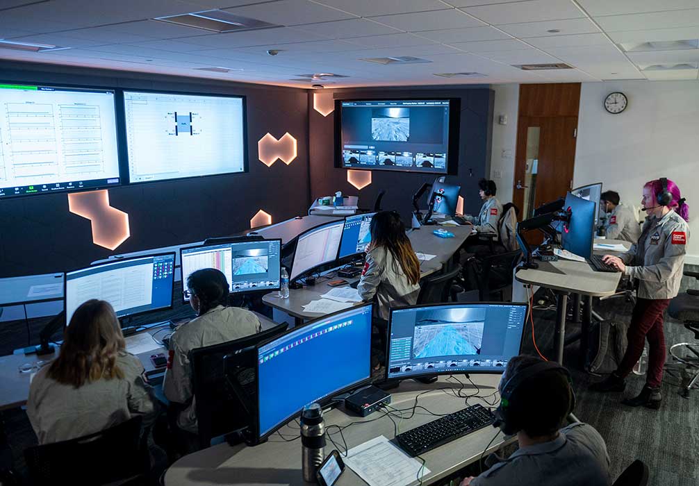 Students sit at large screens in a room with two larger screens in front.