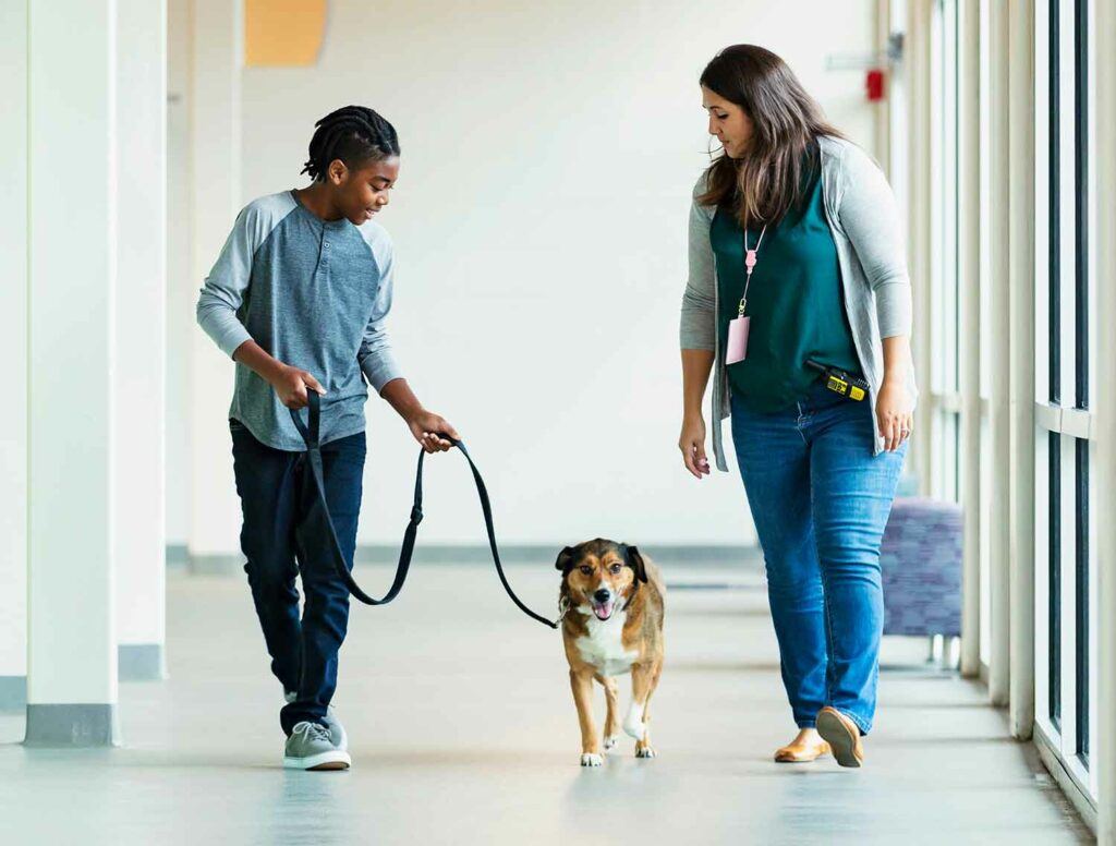A tween child smiles and walks a dog down a hallway and a woman walks with them.