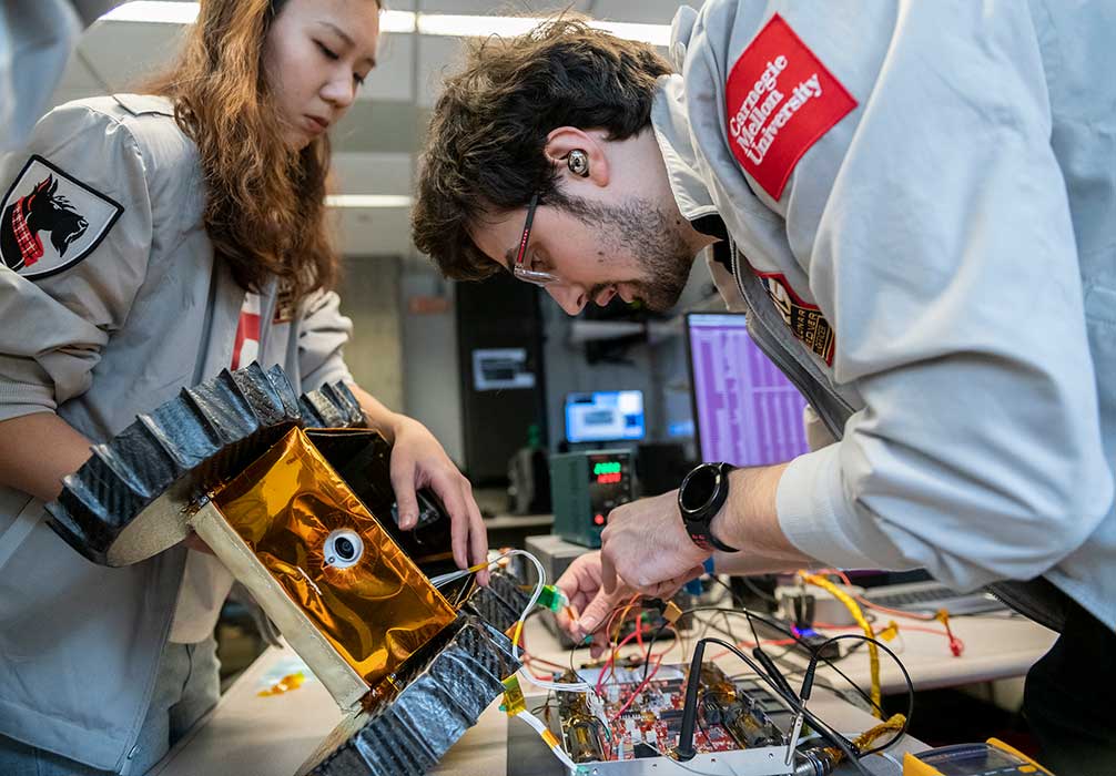 A student holds the top portion of a disassembled rover as another student checks the wires inside.