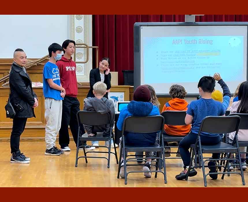 Three teens and an adult stand in front of seated children and next to a screen showing the word AAPI Youth Rising and some information.