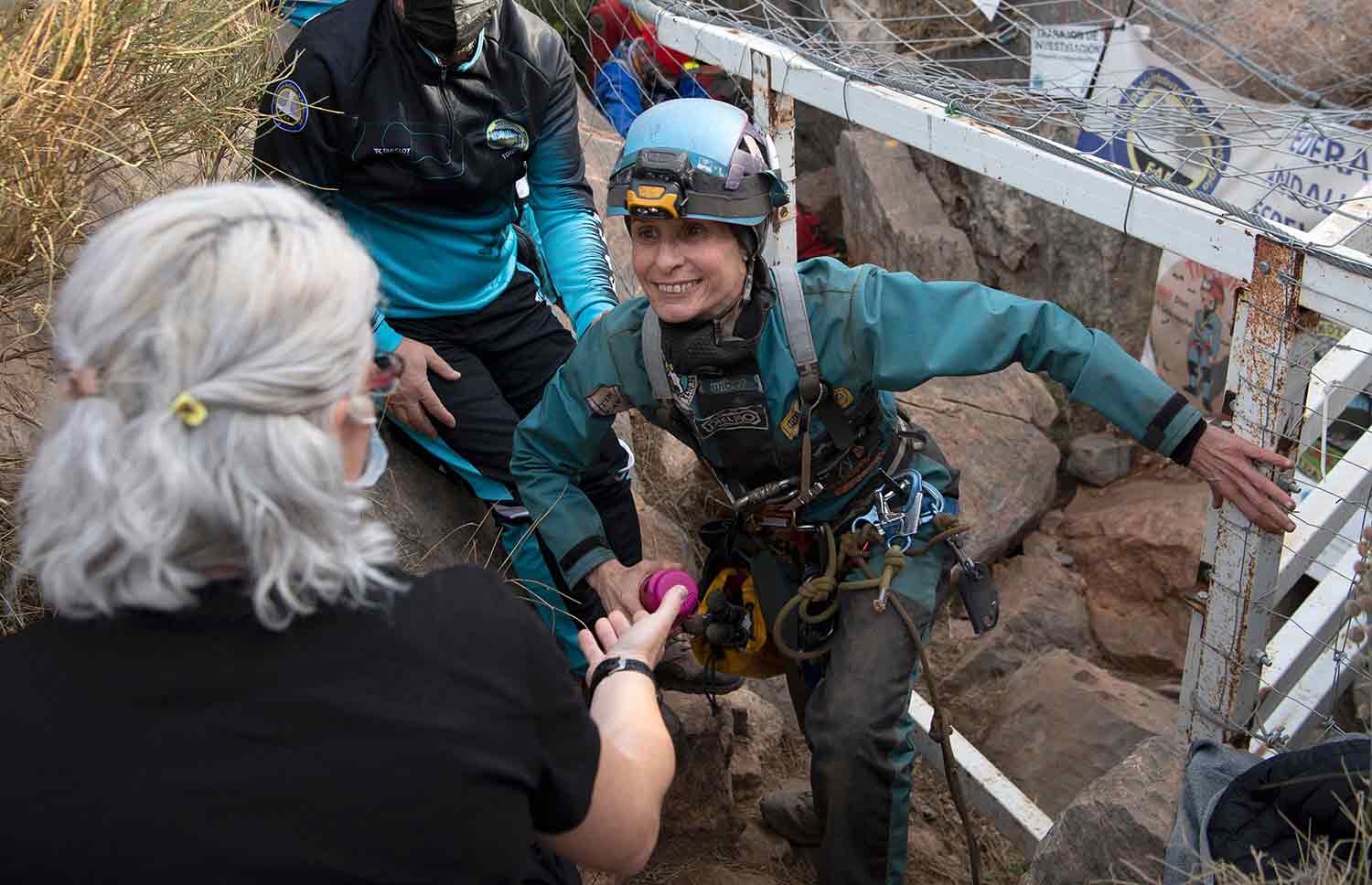 A woman smiles as she walks out of a rock-filled cave as another woman reaches out to assist her.