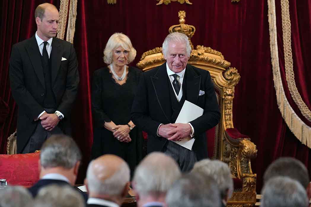 King Charles, Queen Camilla, and Prince William stand before a crowd of people and in front of an ornate gold chair.