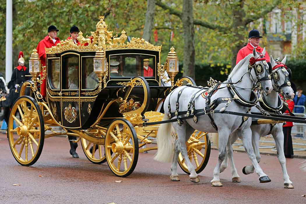 An ornate black and gold coach pulled by two white horses travels down a street.