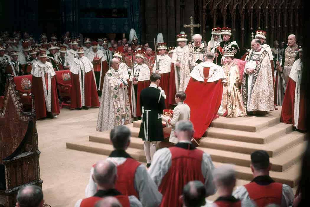 Young Queen Elizabeth sits in a chair wearing a crown and surrounded by people in robes and carrying scepters.
