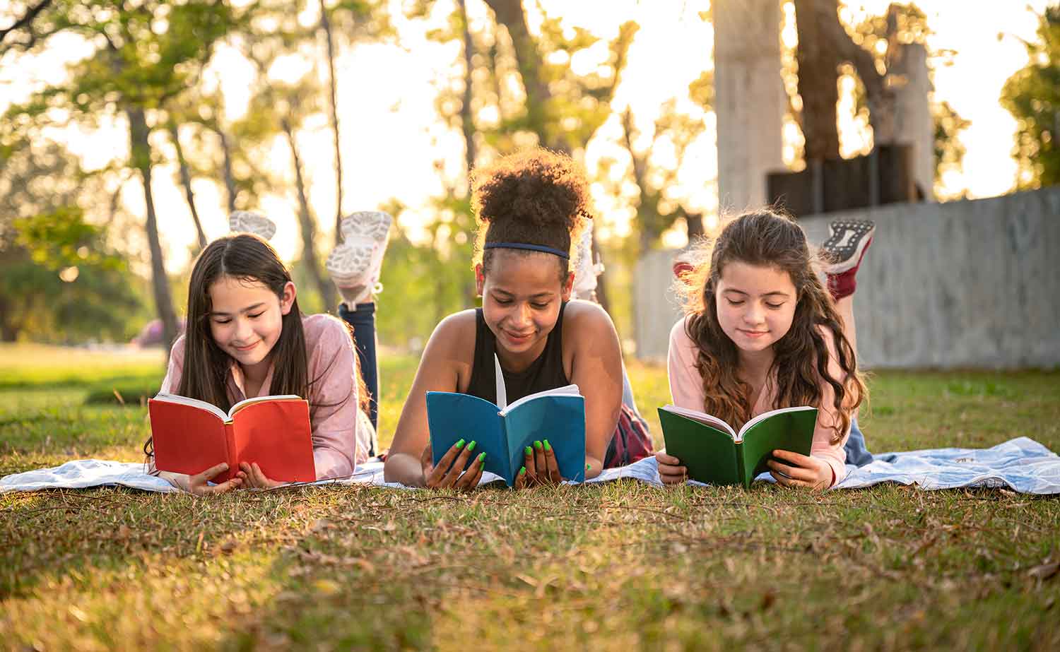 Three girls lie on their stomachs on grass reading books.