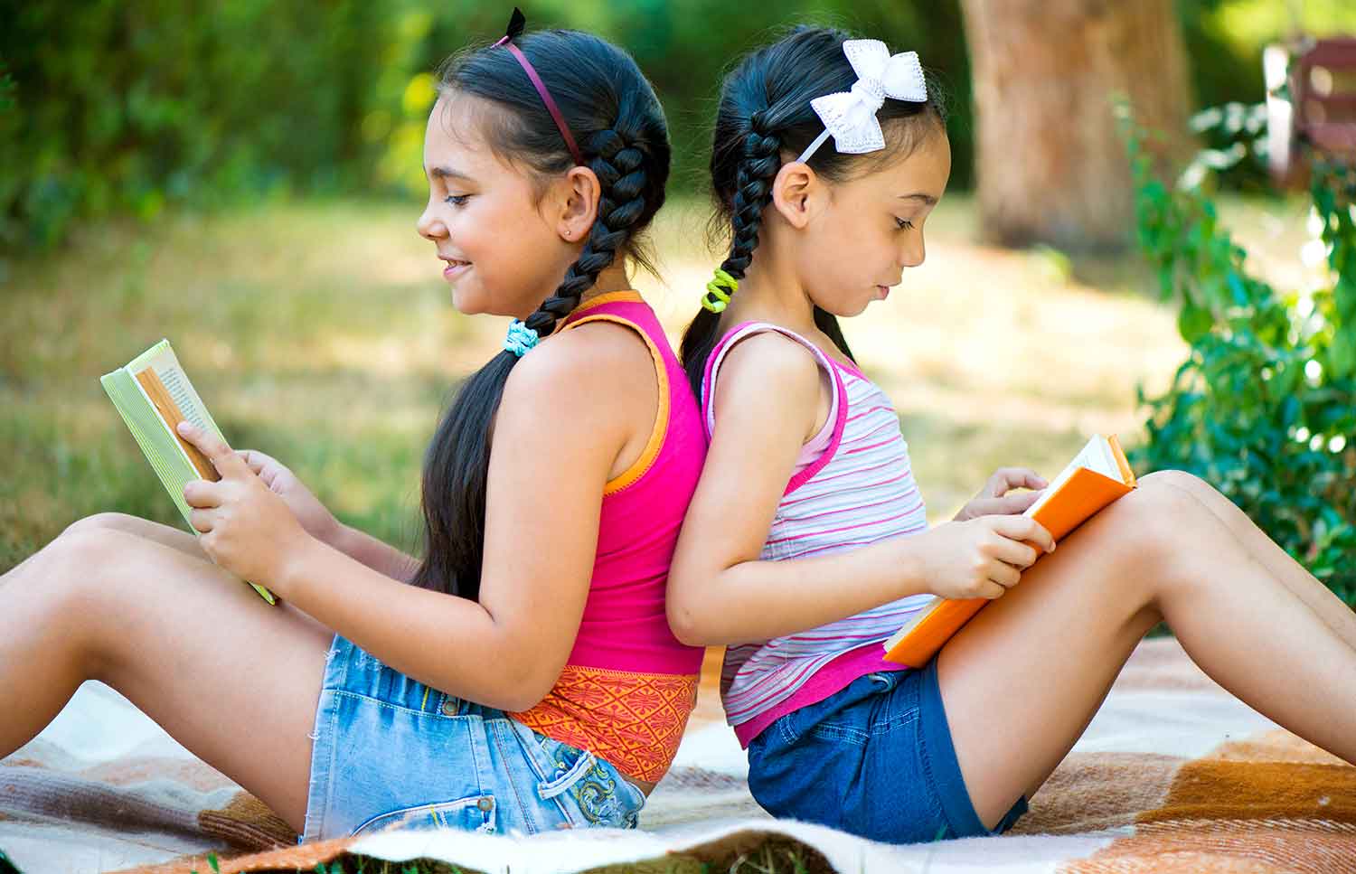 Two girls sit back to back on a blanket outdoors reading books.