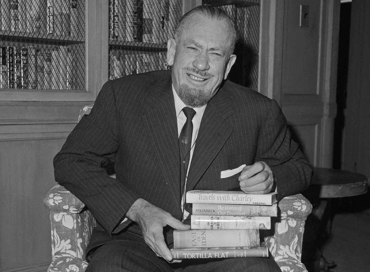 John Steinbeck sits in a chair with a stack of his books on his lap.