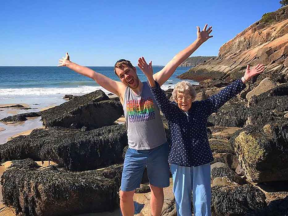 An older woman and a younger man stand on a cliffside beach with their arms outstretched.