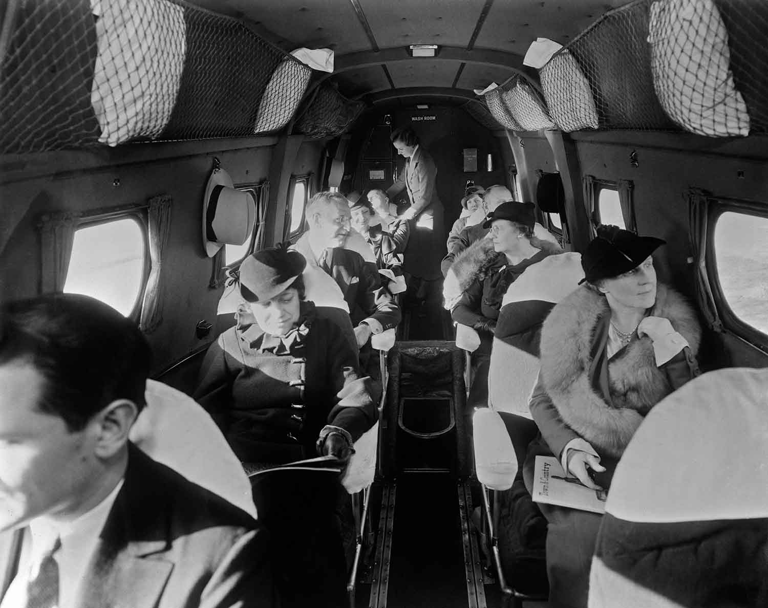 People dressed in 1930s clothing sit in a narrow passenger plane while a flight attendant stands in the aisle.