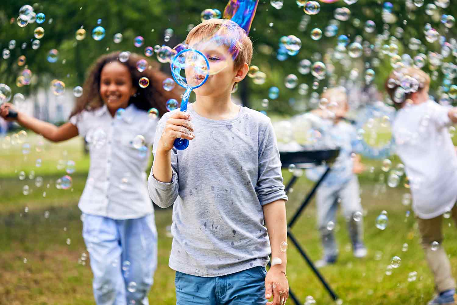 A boy blows bubbles through a wand as kids in the background run through bubbles.