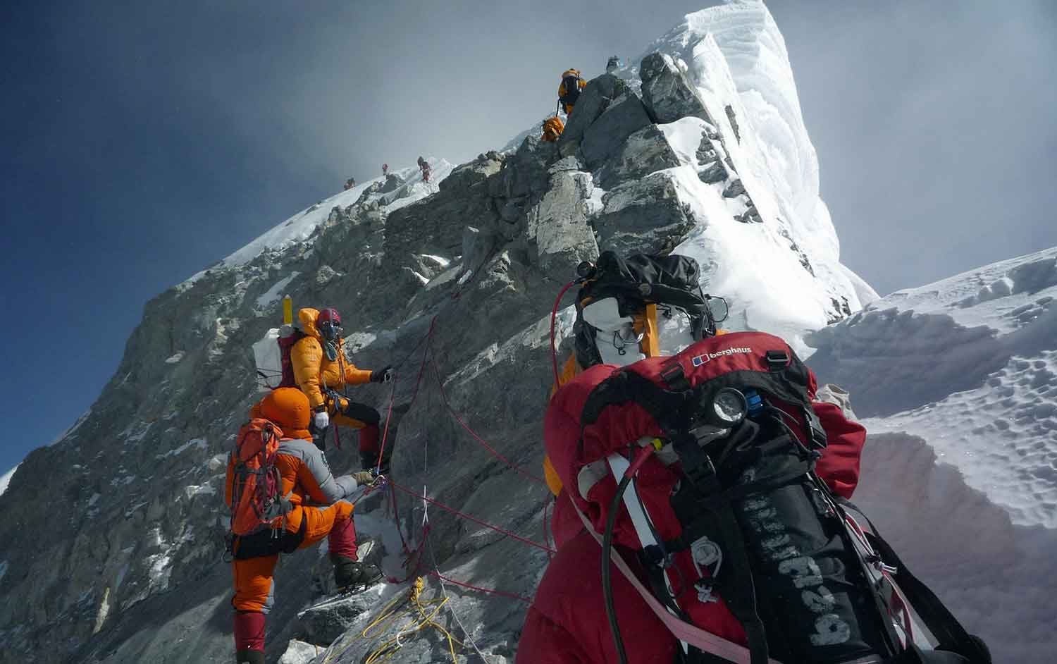 Several people with ropes, oxygen tanks, and other gear climb up the side of a mountain.