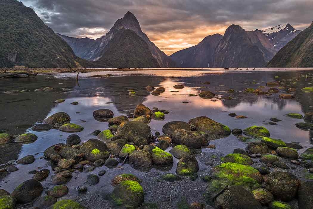 Moss-covered rocks on a body of water with narrow mountains in the background against a sunset.