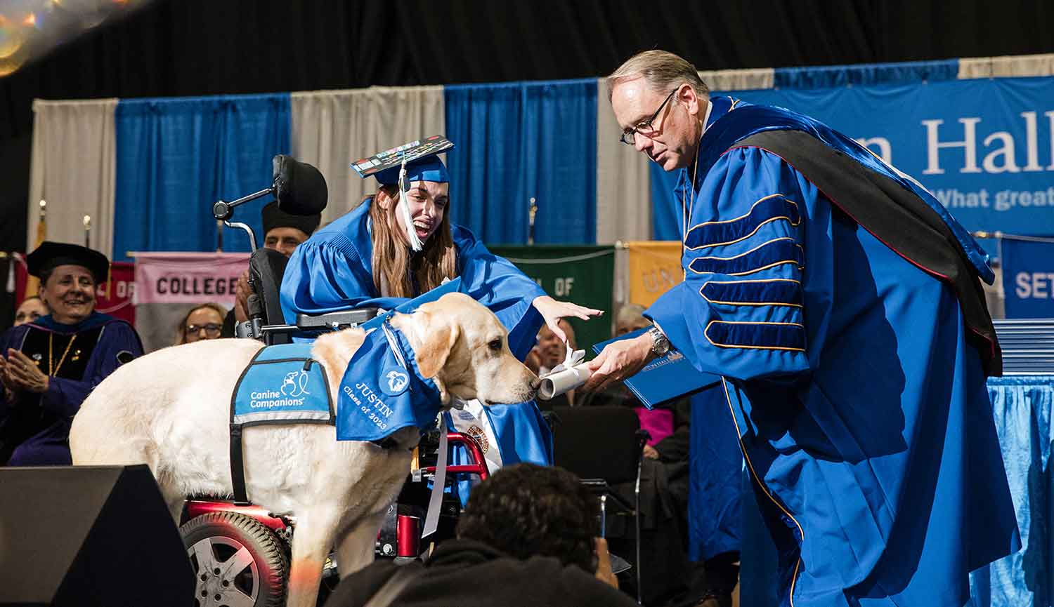 A golden retriever and lab mix dog in a Canine Companions vest sniffs a diploma held out by a man in an academic robe as a woman sitting in a wheelchair and wearing a cap and gown smiles.