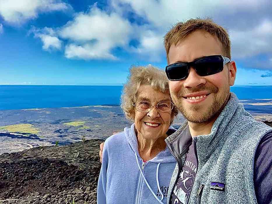 Closeup of an older woman and a younger man smiling in front of a rocky beach and a body of water.