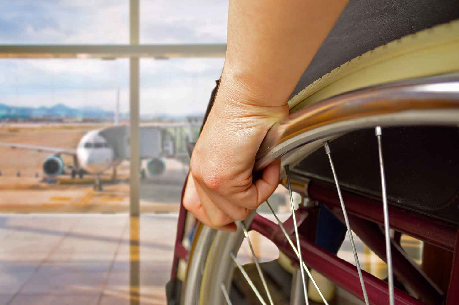Closeup of a hand on the wheel of a wheelchair facing an airport window through which a plane is ready for boarding.