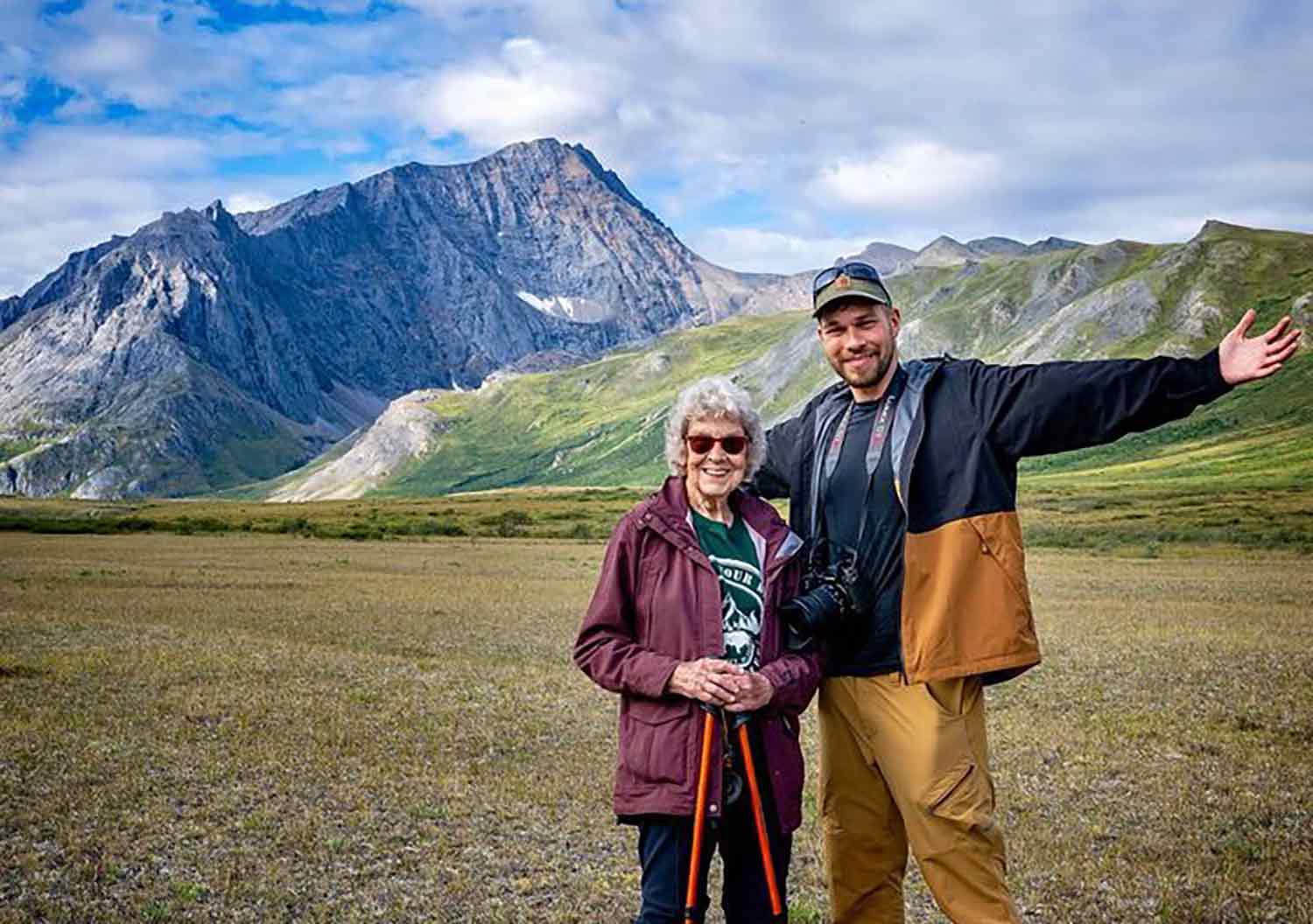 An older woman smiles and poses next to a younger man with his arms outstretched in front of mountains.