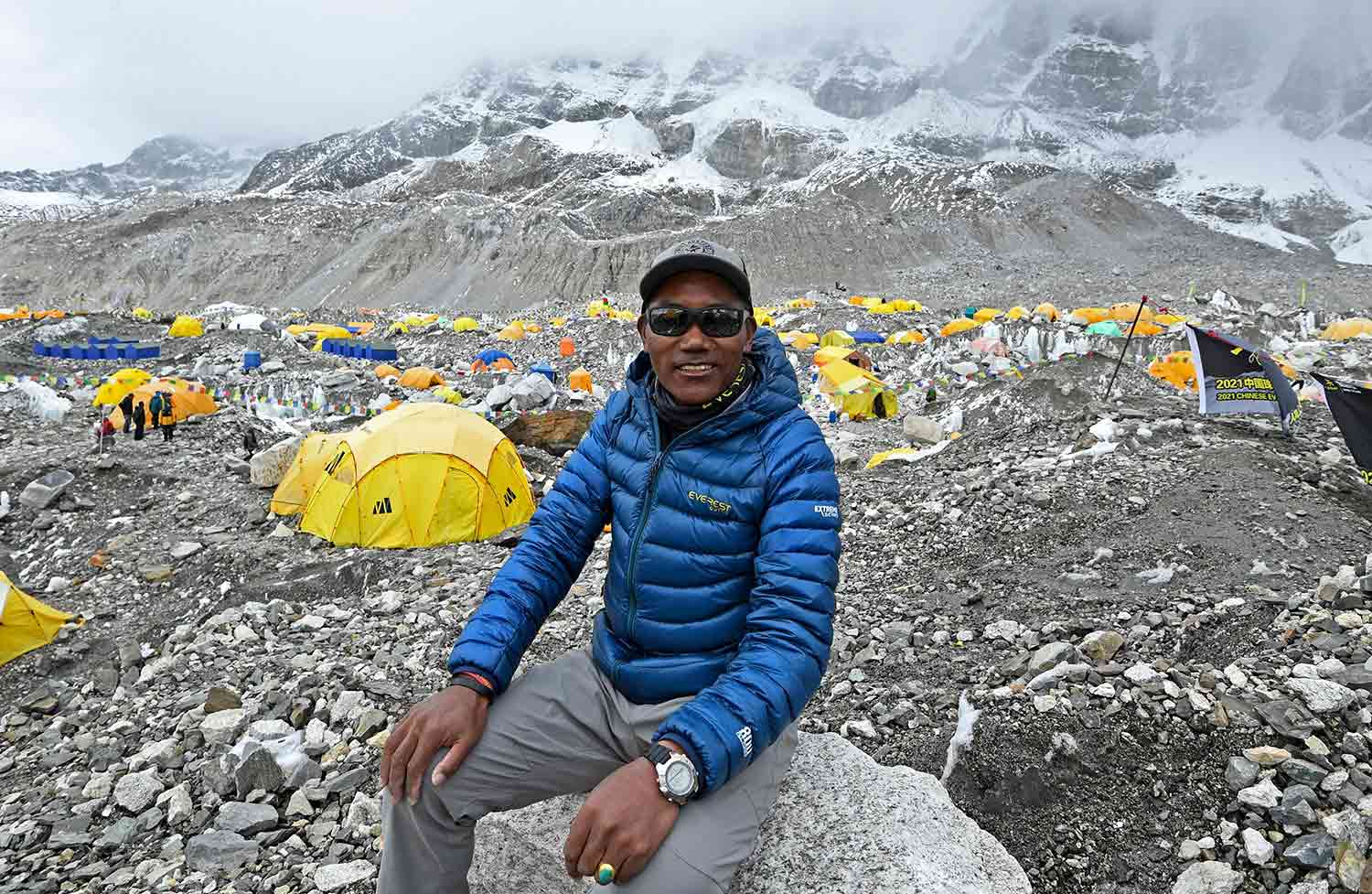 Kami Rita sits and poses on a boulder at Everest base camp with many tents in the background.