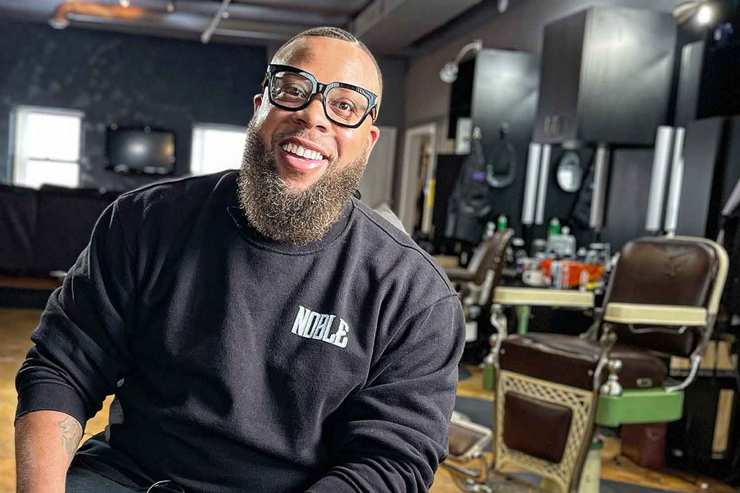 Seated Vernon Jackson smiles with his barber shop in the background.